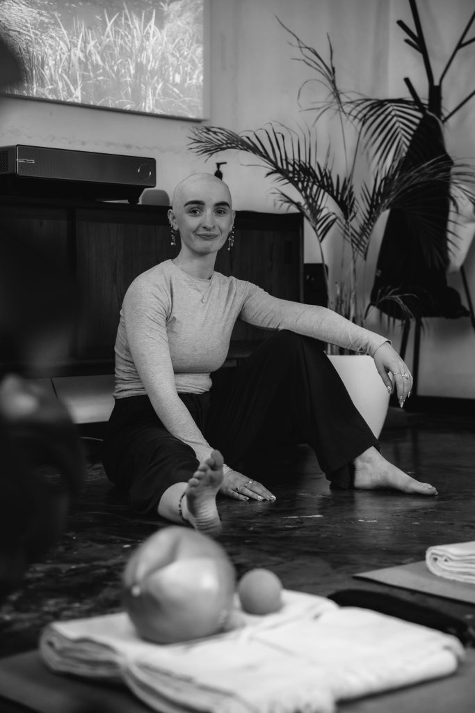 bald woman sitting in a yoga studio, plants, pilates ball, yoga blanket, yoga strap - black and white